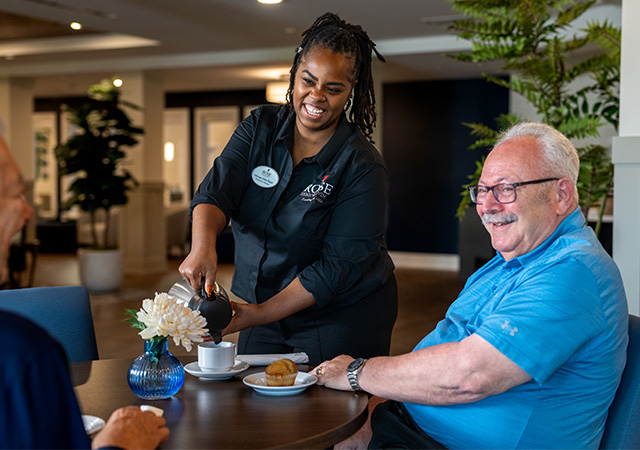 Friendly staff serving coffee to smiling residents in a senior community dining area.
