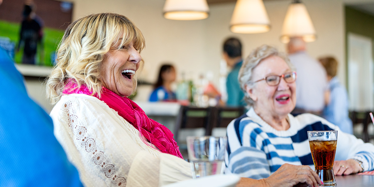 Two elderly women enjoying refreshments in a communal living space with others in the background.