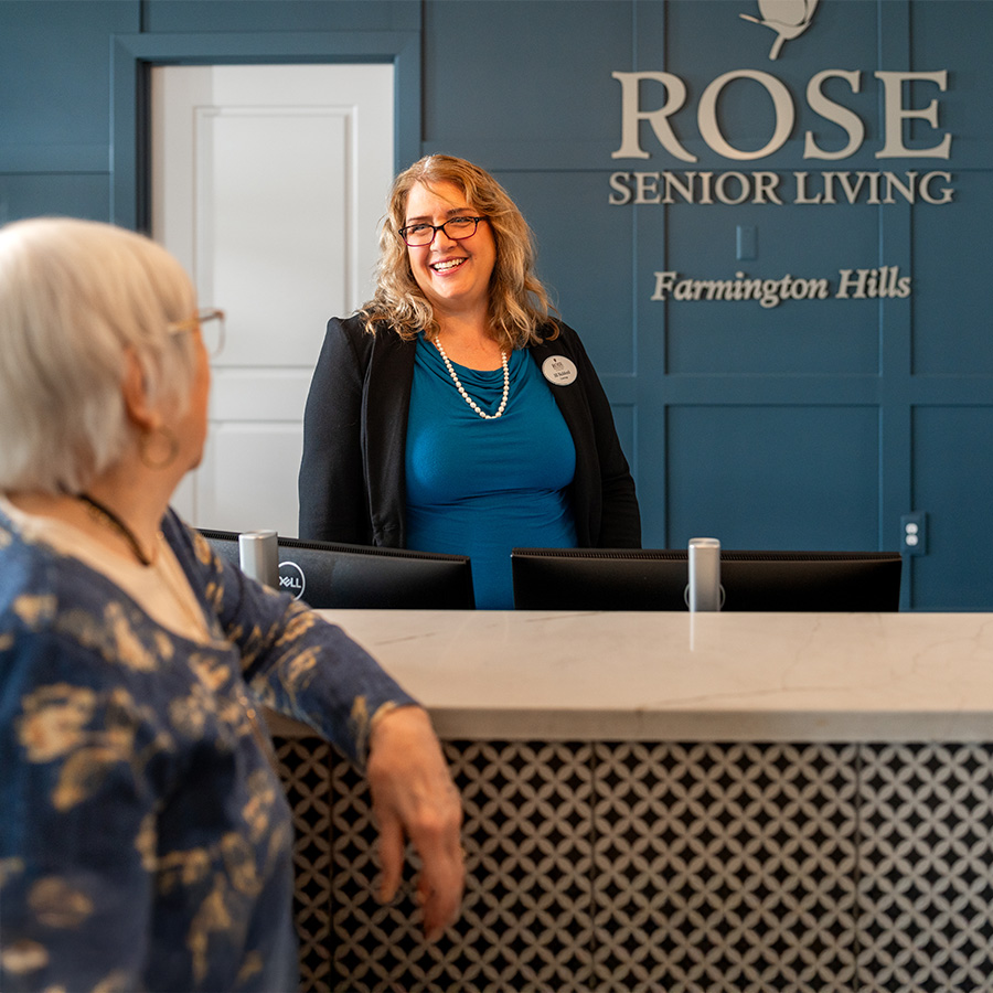 Staff member greeting resident at reception in senior living community.