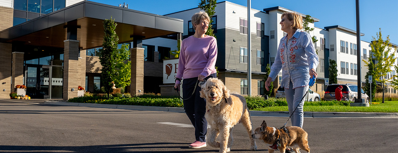 Two women walking dogs in front of a modern residential community building.