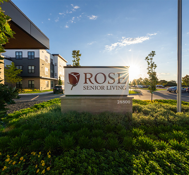 Entrance sign of Rose Senior Living with sunlit building and parking lot in the background.