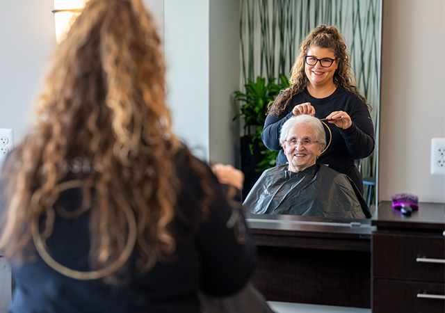 A stylist assists a smiling elderly woman in a hair salon at a senior living community.