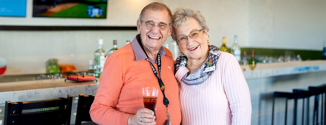 Elderly couple smiling and holding drinks in a communal area of a senior living community.